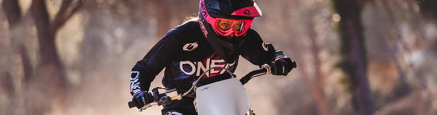 Eine Person mit einem rosa Helm und schwarzer Motocross-Ausrüstung fährt mit einem Dirtbike durch einen staubigen Waldweg.