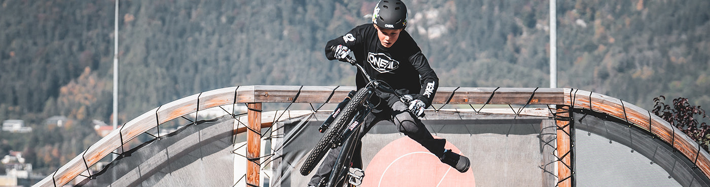 Ein Radfahrer mit Helm und Schutzausrüstung führt auf einer Rampe in einem Skatepark einen Trick vor. Im Hintergrund sind eine Berglandschaft und einige Gebäude zu sehen.