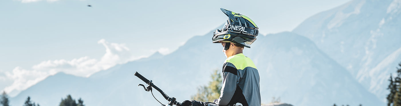 Eine Person mit Helm und Schutzausrüstung steht mit einem Mountainbike da und blickt auf die Berglandschaft im Hintergrund. Der klare blaue Himmel und vereinzelte Wolken bereichern die Landschaft.