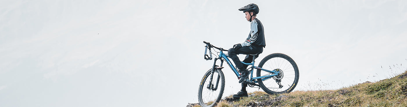 Eine Person mit Helm sitzt auf einem Mountainbike, hält am Rand eines grasbewachsenen Hügels und blickt unter einem bewölkten Himmel in die Ferne.