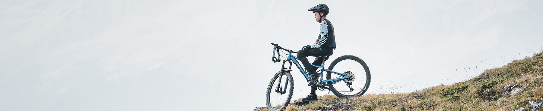 Eine Person mit Helm sitzt auf einem Mountainbike, hält am Rand eines grasbewachsenen Hügels und blickt unter einem bewölkten Himmel in die Ferne.