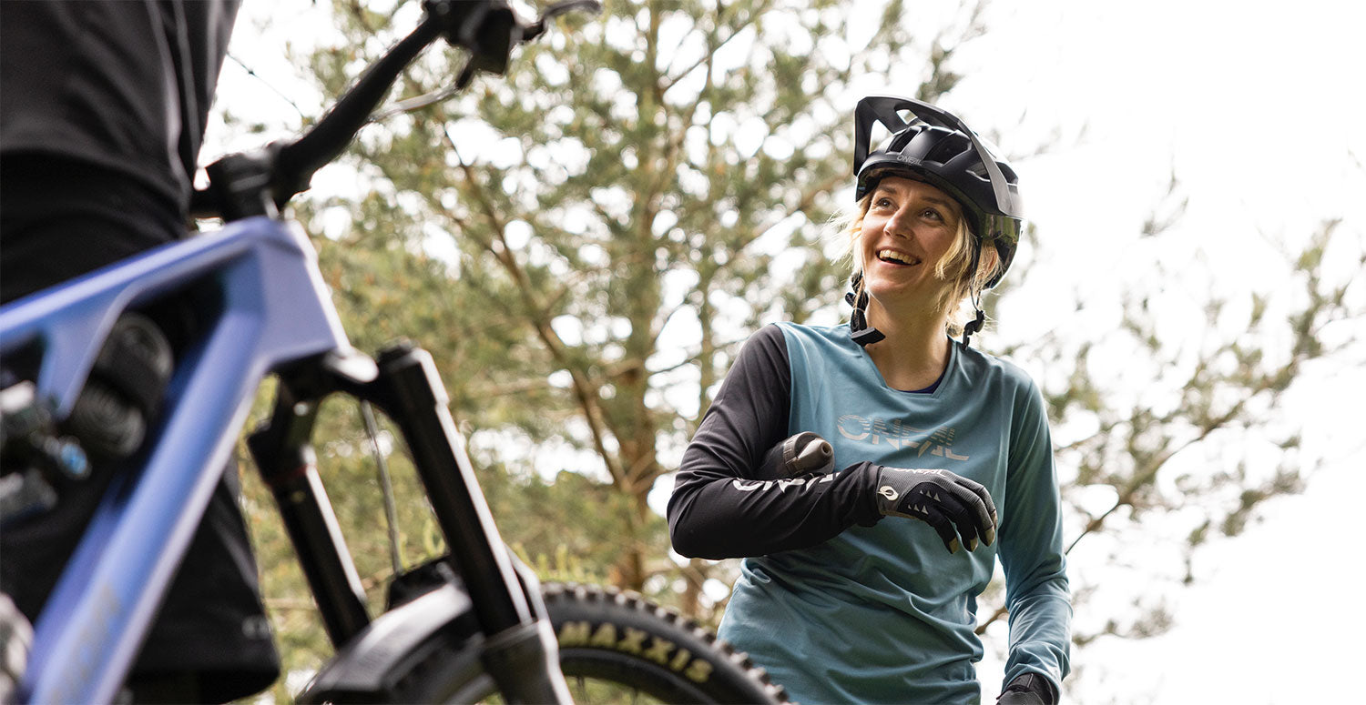 Eine Person mit Helm und blaugrünem Langarmshirt hält inne und lächelt, während sie in einem Waldgebiet Mountainbike fährt. Im Vordergrund ist ein anderer Radfahrer zu sehen, dessen Fahrrad und ein Teil seines Körpers zu sehen sind. Im Hintergrund sind Bäume mit grünem Laub zu sehen.