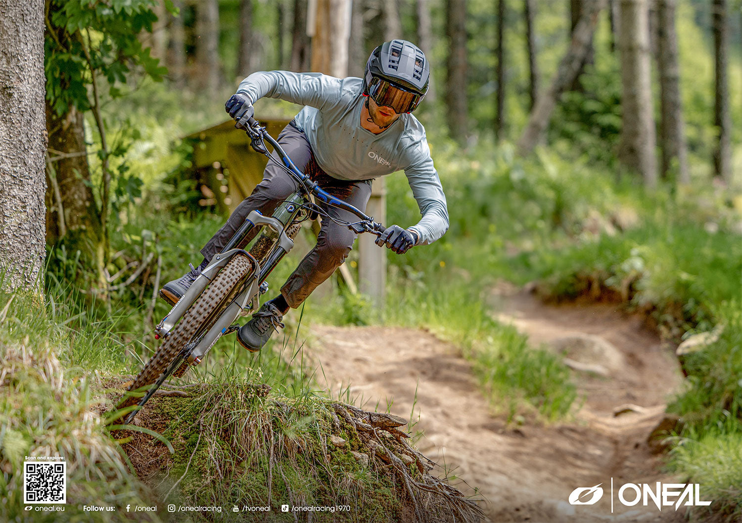 Ein Mountainbiker mit Helm und Schutzausrüstung fährt durch einen Waldweg. Er legt sich in eine Kurve, hält den Lenker fest umklammert und ist von Grün und einem Feldweg umgeben. In der Ecke sind O'NEAL-Markenzeichen und Social-Media-Symbole zu sehen.