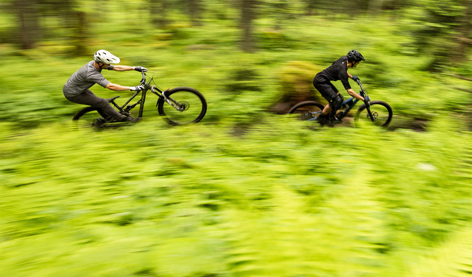 Zwei Mountainbiker fahren mit hoher Geschwindigkeit durch einen üppigen, grünen Waldweg. Die Bewegungsunschärfe betont ihre schnelle Bewegung. Beide tragen Helme und Fahrradausrüstung und navigieren durch das dichte Laub.