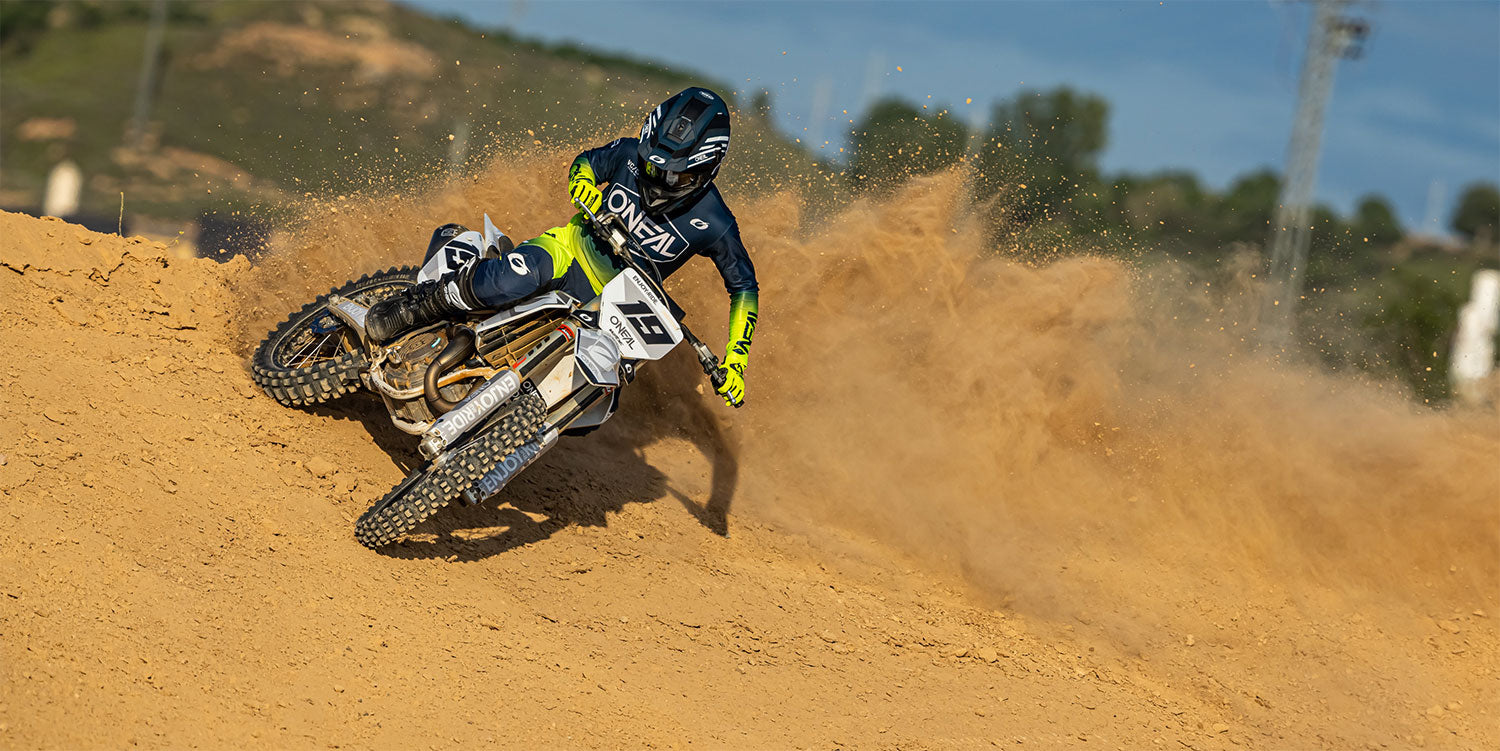 Motocrossfahrer in schwarz-gelber Ausrüstung fährt mit einem weißen Dirtbike über eine staubige Strecke und wirbelt dabei jede Menge Sand auf. Im Hintergrund sind verschwommene Bäume und Hügel unter einem klaren blauen Himmel zu sehen.