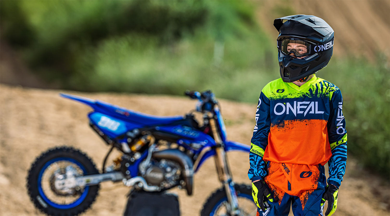 Ein junger Motocrossfahrer in einem orange-blauen O'Neal-Outfit und mit schwarzem Helm steht im Freien neben einem blauen Dirtbike. Im Hintergrund ist ein verschwommenes Naturgelände mit grünem Laub zu sehen. Der Fahrer scheint in die Ferne zu blicken.