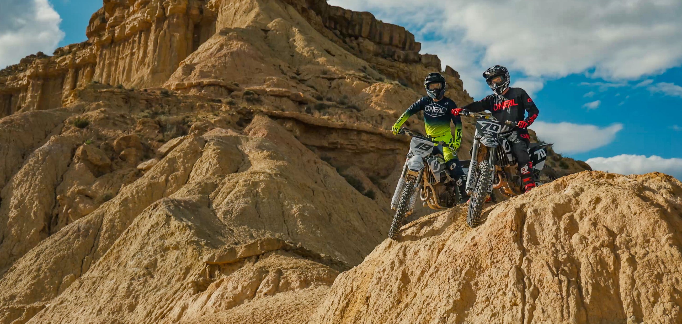 Zwei Dirtbiker in Schutzkleidung, einer in Grün, der andere in Schwarz und Rot, stehen mit ihren Motorrädern auf felsigem Gelände. Sie blicken auf eine ausgedehnte Wüstenlandschaft mit hoch aufragenden Klippen unter einem blauen Himmel mit vereinzelten Wolken.