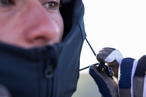 Eine Nahaufnahme zeigt eine Person, die eine O'NEAL CYCLONE Soft Shell Jacke in Blau trägt und den Kordelzug einstellt. Das atmungsaktive Design umfasst Handschuhe mit blauen und weißen Akzenten. Das Foto konzentriert sich auf ihr Auge und einen Teil ihres Gesichts.