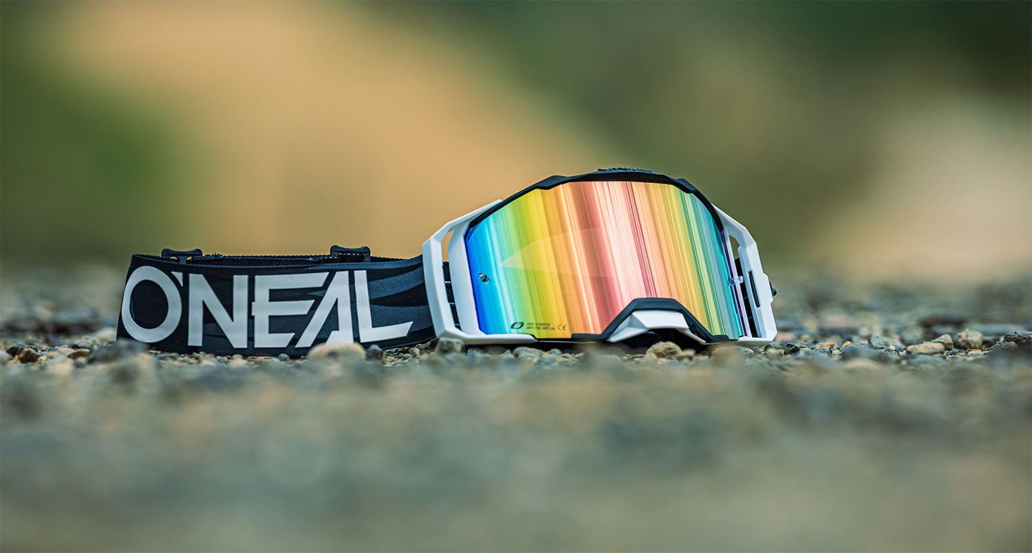 Eine Motocross-Brille mit dem Logo von O'Neal und regenbogenfarbenen Gläsern liegt auf einer felsigen Oberfläche. Das schwarze Band mit dem O'Neal-Logo bildet einen Kontrast zu den reflektierenden, bunten Gläsern. Der Hintergrund ist verschwommen und hebt die Brille hervor.